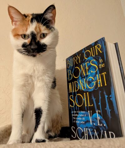 The book, "Bury Our Bones in the Midnight Soil" is standing next to a small calico cat.
