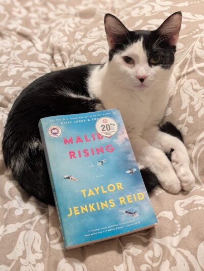 The book, "Malibu Rising" by Taylor Jenkins Reid is lying on a black and white cat that is curled up on a blanket.