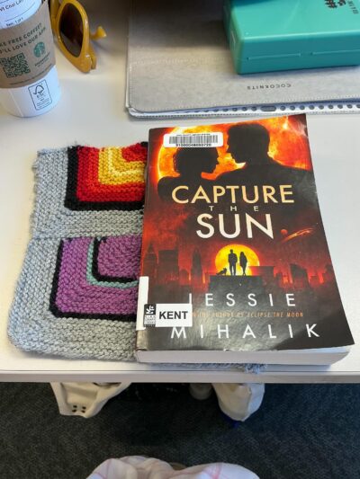 knitted mitered square with grey border and stripes of black, red, orange, and yellow next to a paperback book with the colors black, red, orange, and yellow on the cover. Both sitting on a white table.